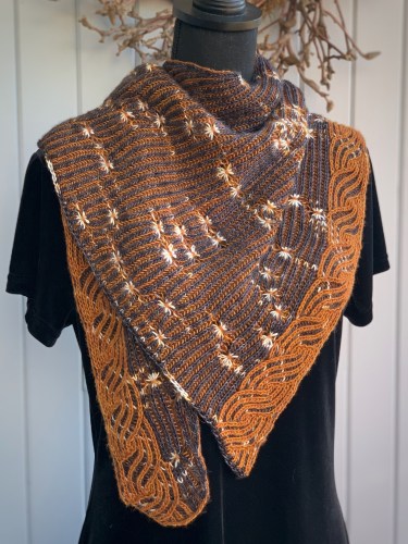 A gray and white brioche knit shawlette featuring assigned pooling stars and a syncopated caramel colored edging