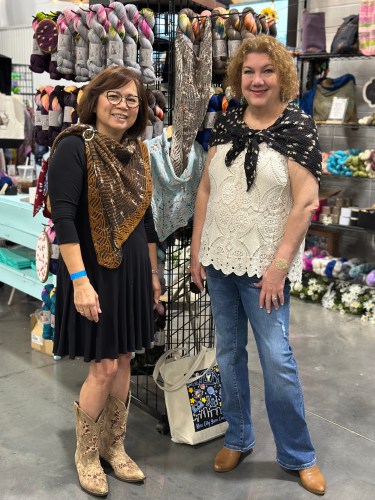 Two women wearing knitted shawls, standing in front of a display of yarn
