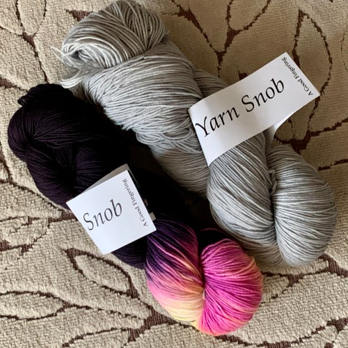 Two skeins of yarn. One is very dark with a pink color pop, and the other is a light gray
